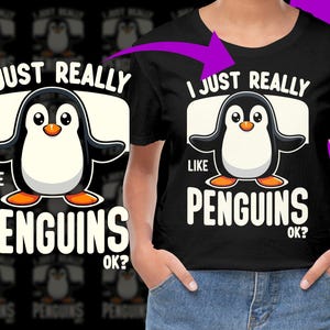May include: Black t-shirt with a cartoon penguin graphic. The penguin is black and white with an orange beak and feet. Text on the shirt reads "I JUST REALLY LIKE PENGUINS OK?" in white block letters.