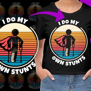Puede incluir: Una camiseta negra con un gráfico de puesta de sol retro y el texto "I do my own stunts". Una figura de dibujos animados con una capa y muletas está en el centro del gráfico.