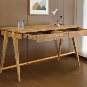 May include: A wooden desk with two drawers, a lamp, and a picture frame on a brown wall. The desk has a light wood finish and a simple design.