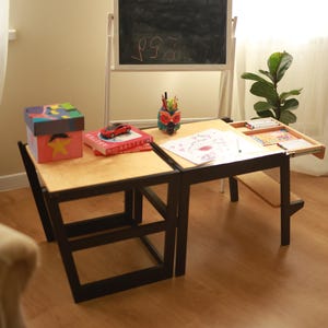 May include: A child's wooden desk with a black frame and a light wood tabletop. The desk has a pull-out tray and is paired with a matching chair. A chalkboard, books, and art supplies are on the desk.