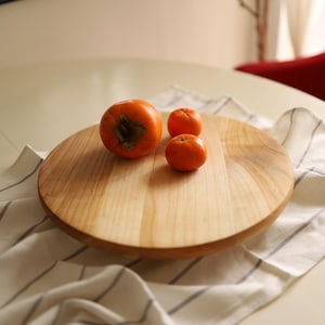 Puede incluir: Una bandeja redonda de madera para servir con tres frutas naranjas en la parte superior. La bandeja es de madera marrón claro y tiene una superficie lisa. Las frutas están dispuestas en línea, con la fruta más grande en el centro.