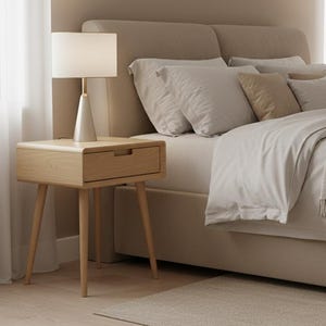 May include: A light wood nightstand with a single drawer and four tapered legs, topped with a conical base lamp featuring a white drum shade. The nightstand sits beside a beige upholstered bed with layered pillows and a textured throw blanket.