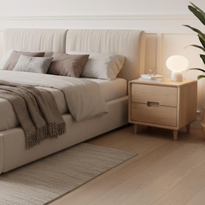 May include: A neutral-toned bedroom scene featuring a bed with a beige headboard and bedding. A brown throw blanket rests on the bed. Two wooden nightstands with lamps flank the bed. A rug and hardwood floor complete the room.