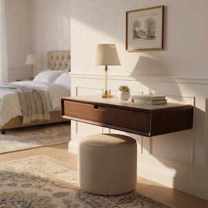 May include: A modern bedroom scene featuring a floating wooden desk with a drawer, a small lamp, and a framed picture. A beige upholstered ottoman sits below the desk. A bed with white linens and a patterned rug complete the room's decor.