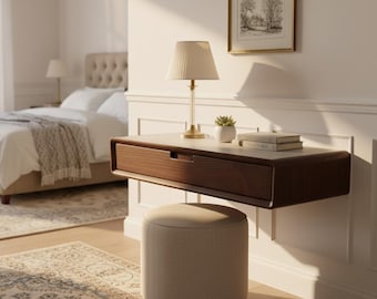 May include: A wall-mounted floating desk with a dark wood drawer, paired with a beige cylindrical ottoman. On the desk sits a table lamp with a cream-colored pleated shade, a small potted succulent, and a stack of two books.