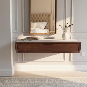 May include: A floating wooden console table with a drawer, mounted on a white wall. A rectangular mirror hangs above the table, reflecting a bed. A small white vase with greenery and a stack of books are on the table.