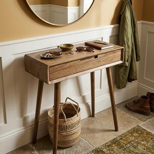 Peut inclure: Une console rectangulaire en bois dotée d'un tiroir unique et de quatre pieds fuselés. Le plateau supporte une petite coupelle avec des clés, un bol, des lunettes de soleil et un carnet. Un panier tressé est posé au sol sous la table en bois.