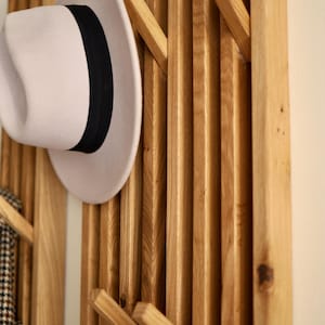 May include: A wall-mounted coat rack constructed from vertical light brown wooden slats with integrated angled pegs. A light beige fedora hat with a black ribbon band is hanging on one of the pegs, and a patterned fabric item hangs on another.