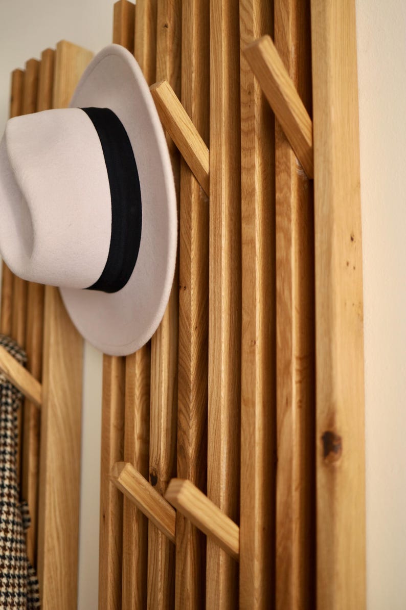 May include: A wall-mounted coat rack constructed from vertical light brown wooden slats with integrated angled pegs. A light beige fedora hat with a black ribbon band is hanging on one of the pegs, and a patterned fabric item hangs on another.