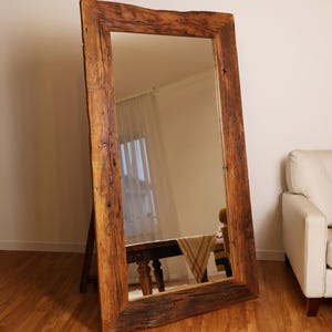 Può includere: Uno specchio grande con cornice in legno rustico di forma rettangolare. La cornice è realizzata in legno riciclato con una finitura marrone naturale. Lo specchio è appoggiato a una parete in una stanza con pavimento in legno.
