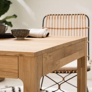 Peut inclure: Table de salle à manger en bois clair avec un tiroir visible. Un bol à motifs et un tissu blanc roulé sont posés sur la table. Une chaise en osier est en arrière-plan, suggérant un cadre de salle à manger.
