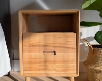 Puede incluir: Una mesita de noche de madera clara con un estante superior abierto y dos cajones con tiradores horizontales integrados. El mueble se apoya sobre cuatro patas de madera cónicas y muestra un patrón de veta de madera natural visible.