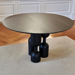 May include: A round, dark wood table with a unique base design. The tabletop is a light gray color, and the base is black with a sculptural, rounded shape. The table is set on a light-colored rug.