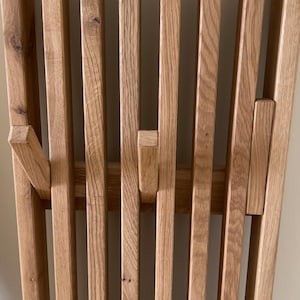 May include: A wall-mounted coat rack constructed from vertical light brown oak wood slats. The design features integrated, retractable hooks that fold out from the slatted structure, showcasing the natural wood grain and texture of the timber.