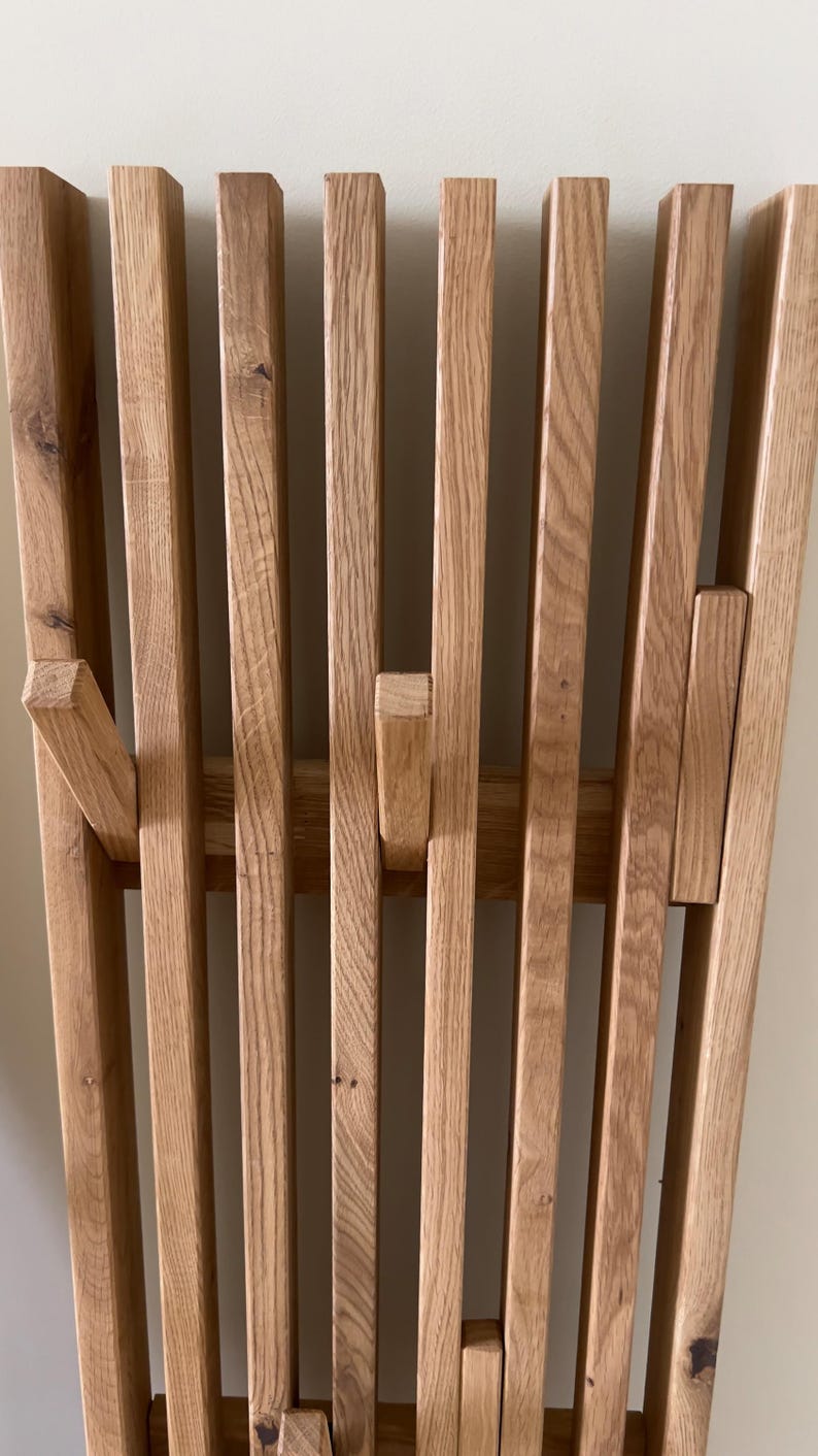 May include: A wall-mounted coat rack constructed from vertical light brown oak wood slats. The design features integrated, retractable hooks that fold out from the slatted structure, showcasing the natural wood grain and texture of the timber.