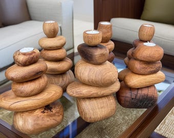 May include: Three decorative candle holders crafted from stacked, smooth, natural-toned wooden stones. Each sculpture features a rounded wooden base with several smaller, polished wooden elements piled vertically, topped with a small white tea light candle.