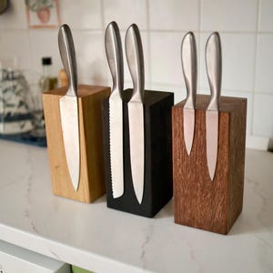 May include: Three wooden knife blocks in varying colors: light, black, and dark brown. Each block holds stainless steel knives. The knives have silver blades and handles. The blocks are rectangular and sit on a white countertop.