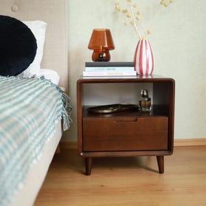 Può includere: Un comodino in legno scuro con un cassetto e un ripiano aperto. Sopra si trovano una lampada a fungo in vetro marrone, due libri impilati e un vaso a righe rosa e bianche con steli secchi. Sul ripiano ci sono un piccolo vassoio e un flacone di profumo.