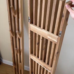 May include: Two vertical wall-mounted coat racks made of light-toned wood. Each rack features a slatted design with multiple protruding wooden pegs for hanging items. The back of the rack shows metal keyhole mounting hardware for installation.