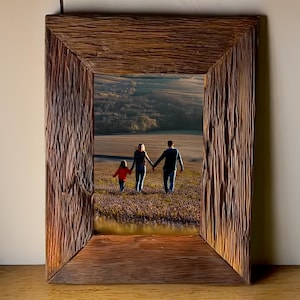May include: A rectangular picture frame crafted from dark brown, weathered wood with a deeply grooved, textured surface. The frame contains a photograph of three people walking hand-in-hand through a field of purple flowers at sunset.