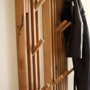 May include: A wall-mounted coat rack constructed from vertical light brown wooden slats with integrated angled pegs. The rack features a natural wood grain texture and is shown with a black leather jacket hanging from one of the upper wooden hooks.
