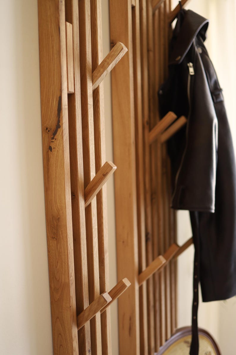 May include: A wall-mounted coat rack constructed from vertical light brown wooden slats with integrated angled pegs. The rack features a natural wood grain texture and is shown with a black leather jacket hanging from one of the upper wooden hooks.