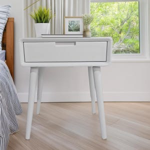 May include: A white nightstand with a single drawer and four angled legs. The nightstand has a minimalist design and is placed next to a bed. A small framed picture and a potted plant are on top of the nightstand.