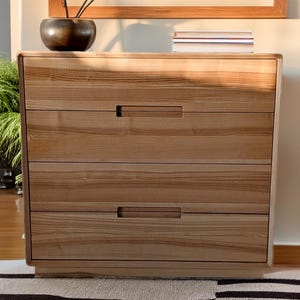 Könnte beinhalten: Eine Kommode aus hellem Holz mit einem rechteckigen Rahmen und drei horizontalen Schubladen. Jede Schublade ist mit einem eingelassenen, integrierten Griff ausgestattet, der mittig an der Oberkante sitzt und eine natürliche Holzmaserung zeigt.