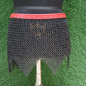 Könnte beinhalten: Ein schwarzer Kettenhemdrock mit einem roten Ledergürtel. Der Rock hat einen spitzen Saum und ein dekoratives Metallemblem auf der Vorderseite.