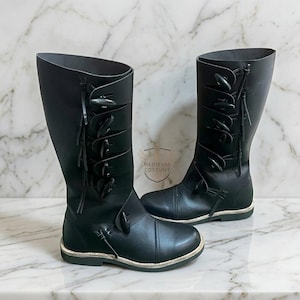 May include: A pair of black leather knee-high boots. The boots have multiple decorative toggles and straps. The boots have a rounded toe and a flat sole. The boots are displayed on a marble surface.