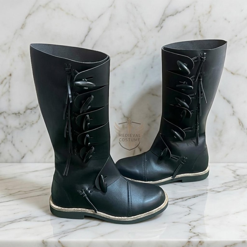 May include: A pair of black leather knee-high boots. The boots have multiple decorative toggles and straps. The boots have a rounded toe and a flat sole. The boots are displayed on a marble surface.