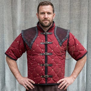 May include: A burgundy quilted medieval gambeson with black leather accents and gold-coloured studs. The short-sleeved garment features a high collar, decorative shoulder armour, and leather straps with buckles. Suitable for historical reenactment or costume.