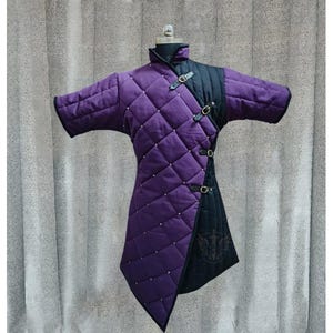 May include: A quilted purple and black medieval gambeson, displayed on a mannequin. The purple section features a diamond pattern with small silver studs. The black section has leather straps with buckles. The garment has short sleeves and a high collar.