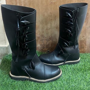 May include: A pair of black leather knee-high boots. Each boot has multiple leather straps with decorative toggles and fringe details. The boots are displayed on a bed of green artificial grass.