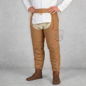 May include: Brown quilted medieval leg armor with a leather belt and ties. The armor is designed to protect the legs and thighs. The image also includes brown boots. The Medieval Costume logo is visible on the right side.