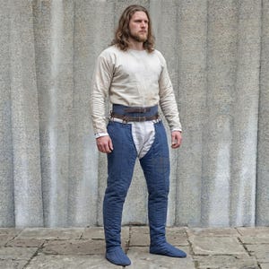 May include: A long-sleeved cream-colored shirt paired with blue quilted leg coverings that feature a high-waisted design. The leg coverings are secured at the waist with a dark brown leather belt featuring two metal buckles and straps.