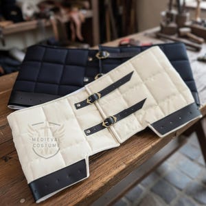 Könnte beinhalten: Zwei gesteppte mittelalterliche Kostümteile, eines cremefarben, das andere schwarz. Beide haben schwarze Lederbesätze, Riemen und Messingschnallen. Das cremefarbene Teil trägt den Text "MEDIEVAL COSTUME" mit einem Wappenlogo. Auf einer Holzoberfläche.