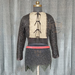 May include: A medieval chainmail shirt and skirt set displayed on a mannequin. The shirt has long sleeves and a beige panel with black ties. A red belt accents the waist, and the skirt is also chainmail.
