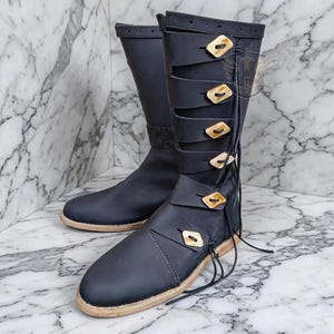 May include: A pair of black leather boots with decorative wooden buttons and straps. The boots have a tall shaft and a light-coloured sole. The boots are displayed on a marble surface.