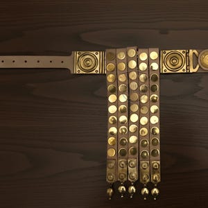 May include: A tan leather belt with brass accents. The belt features decorative brass squares and circular studs. The belt has a series of hanging leather straps with brass studs and decorative brass pendants.