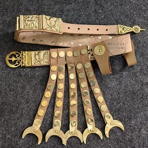 May include: A leather belt with gold-toned metal accents and a matching set of five leather straps with gold-toned metal accents. The belt has a buckle with a design of a Roman soldier. The straps have a crescent-shaped design at the end. The text "MEDIEVAL COSTUME" is visible on the belt.