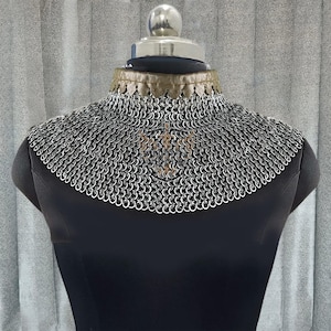 Könnte beinhalten: Ein silberner Kettenkragen, der auf einer schwarzen Schaufensterpuppe ausgestellt ist. Der Kragen besteht aus kleinen, ineinandergreifenden Metallringen, die einen flexiblen, schützenden Nackenschutz bilden. Der obere Rand hat eine dekorative Verzierung.