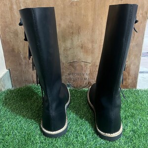 May include: A pair of black leather knee-high boots with decorative tassels and a round toe. The boots have a flat sole with a light-coloured trim. The boots are displayed on green grass.