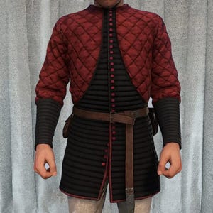 Könnte beinhalten: Ein mittelalterliches Kleidungsstück mit einer gesteppten, rautenförmigen roten Jacke über einem schwarzen, vertikal gestreiften Überrock. Das Outfit umfasst schwarze Ärmel, einen braunen Gürtel und rote Knöpfe. Das Design deutet auf historische oder Fantasy-Kleidung hin.