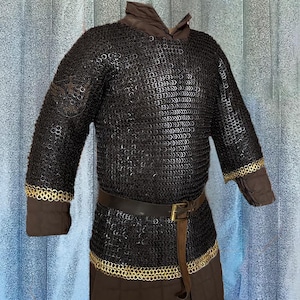 Puede incluir: Una camisa de cota de malla negra con un cinturón de cuero marrón y ribetes dorados. La camisa tiene mangas largas y un escote redondo.