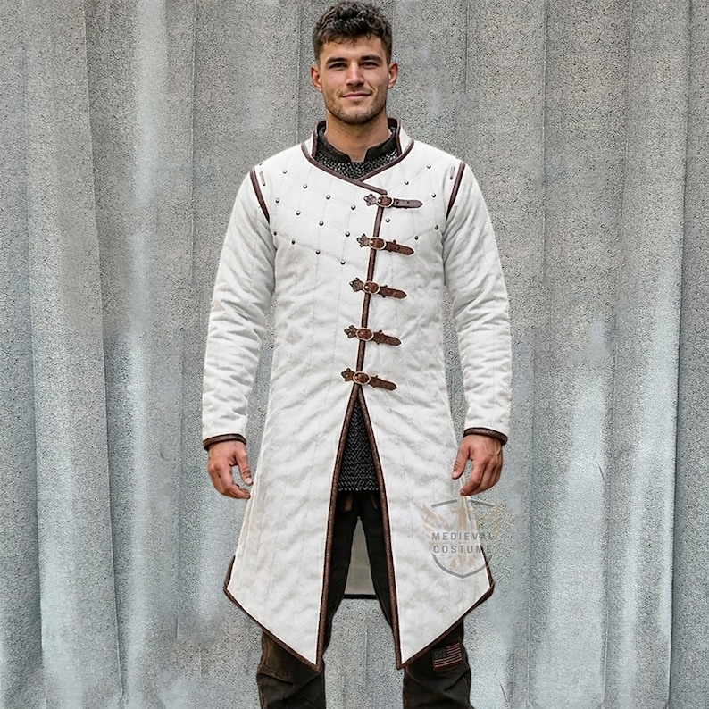 Pu&ograve; includere: Un cappotto medievale bianco a maniche lunghe con finiture e fibbie in pelle marrone. Il cappotto ha un design trapuntato e viene indossato sopra una cotta di maglia. Il cappotto ha il testo "MEDIEVAL COSTUMES".
