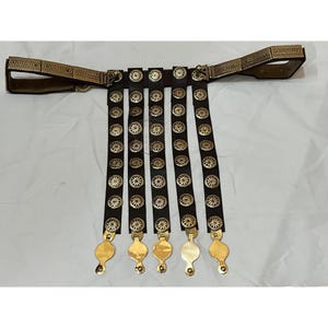 May include: A brown leather belt with five rows of gold-colored metal discs. Each disc has a floral design. The belt has a gold-colored buckle and a gold-colored strap with a decorative design.