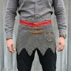 May include: A silver chainmail skirt with a red leather belt and brown leather straps. The skirt has a scalloped hemline and is worn over black pants. The belt has two buckles.