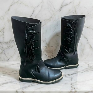 May include: A pair of black leather knee-high boots. The boots have multiple decorative toggles and straps. The boots have a rounded toe and a flat sole. The boots are displayed on a marble surface. The text "MEDIEVAL COSTUME" is visible.