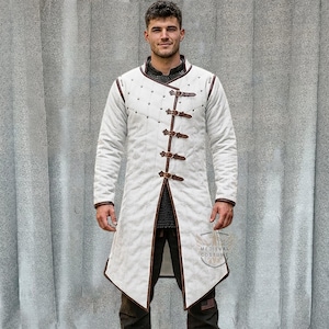 Pu&ograve; includere: Un cappotto medievale bianco a maniche lunghe con finiture e fibbie in pelle marrone. Il cappotto ha un design trapuntato ed &egrave; fissato con diversi cinturini in pelle. Parte di un costume medievale.